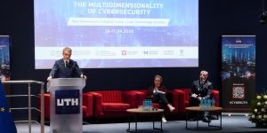 Mężczyzna przemawia na konferencji o wielowymiarowości cyberbezpieczeństwa, a w tle widoczne są czerwone kanapy i prezentacja.