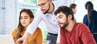 Nauczyciel wskazuje na ekran laptopa, podczas gdy dwójka uczniów uważnie obserwuje, ucząc się razem.