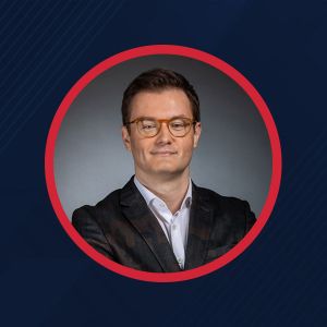 Mężczyzna w okularach z uśmiechem, ubrany w czarną marynarkę, pozujący na tle ciemnoniebieskiego tła.
