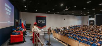 Uczestnicy ceremonii ukończenia studiów UTH w 2026 roku słuchają przemówienia na tle flag i dekoracji.