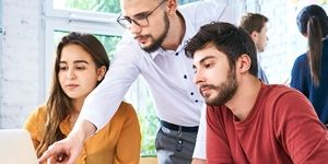 Nauczyciel wskazuje na ekran laptopa, podczas gdy dwójka uczniów uważnie obserwuje, ucząc się razem.