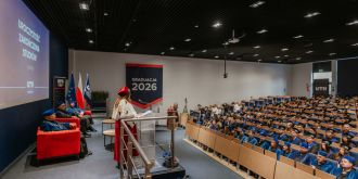Uczestnicy ceremonii ukończenia studiów UTH w 2026 roku słuchają przemówienia na tle flag i dekoracji.
