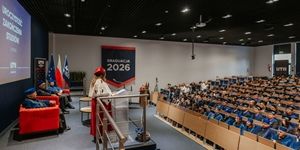 Ceremonia graduacji 2026 w auli, z mówcą przed zgromadzonymi absolwentami i flagami narodowymi.