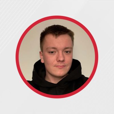 Młody mężczyzna w czarnej bluzie z kapturem, uśmiechający się do aparatu na jasnym tle.