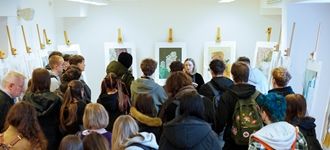 Grupa studentów ogląda wystawę sztuki, słuchając prelekcji prowadzonej przez artystkę.