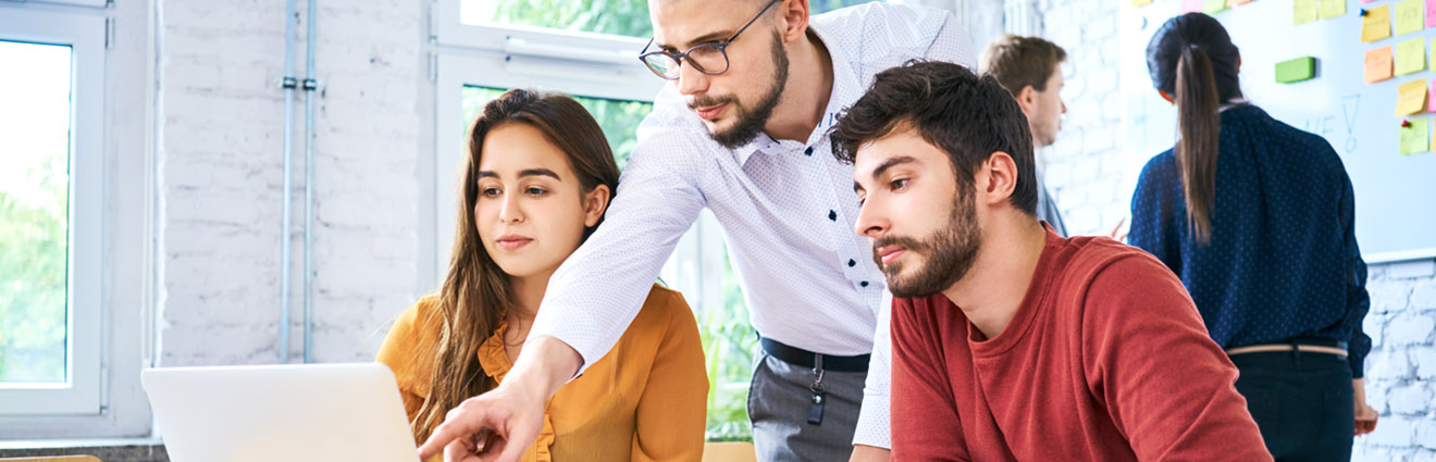 Trzech studentów uczy się wspólnie przy laptopie, podczas gdy nauczyciel wskazuje ważne informacje na ekranie.