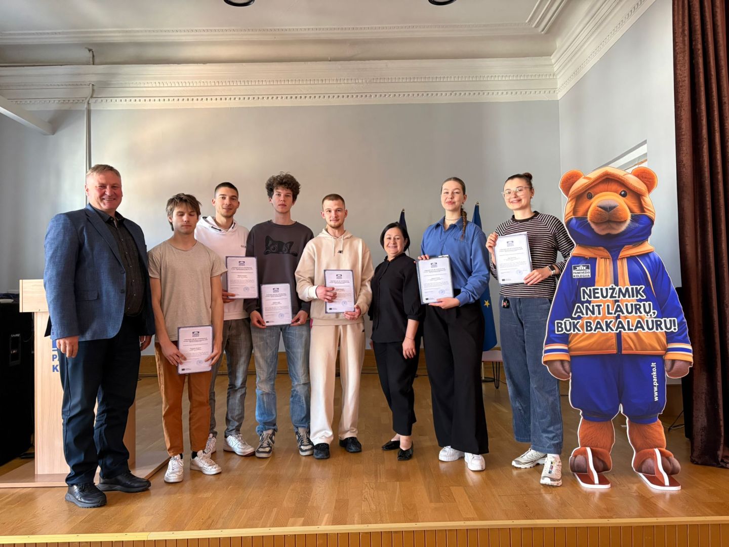 Uczestnicy ceremonii wręczenia dyplomów stoją razem z maskotką, świętując osiągnięcia edukacyjne.