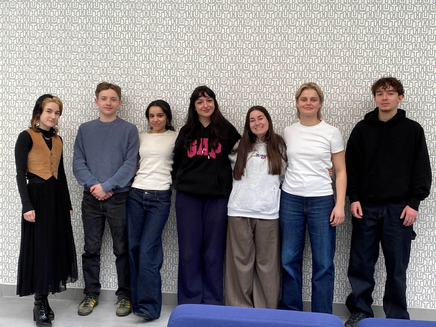 Seven students standing in a row against a patterned wall in an indoor setting.