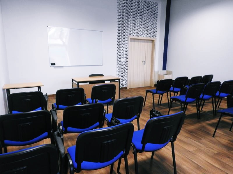 Sala wykładowa z rzędami niebiesko-czarnych krzeseł zwróconych w stronę tablicy i biurka.