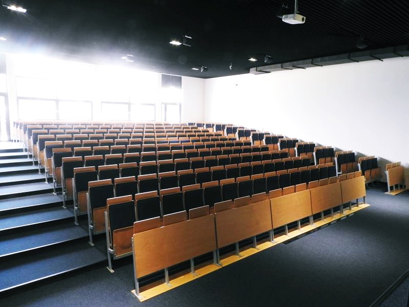 Sala wykładowa z drewnianymi i tapicerowanymi siedzeniami, białymi ścianami i ciemnym sufitem z projektorem.