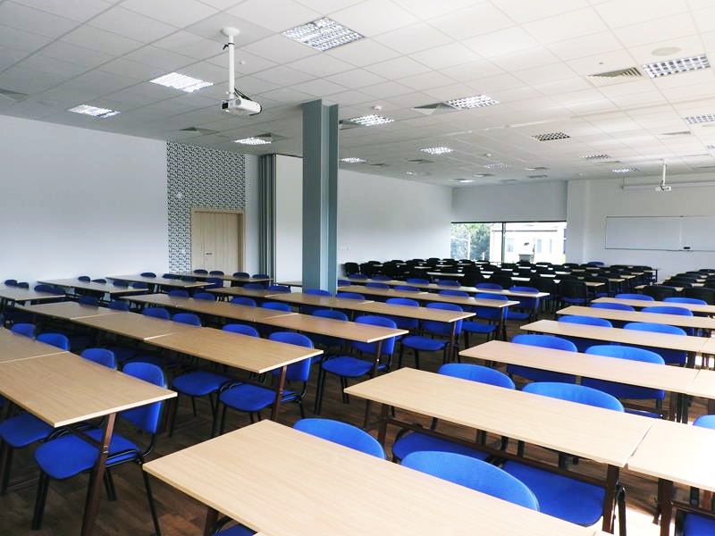 Duża sala wykładowa z rzędami drewnianych stolików, niebieskimi krzesłami, projektorem i tablicą.