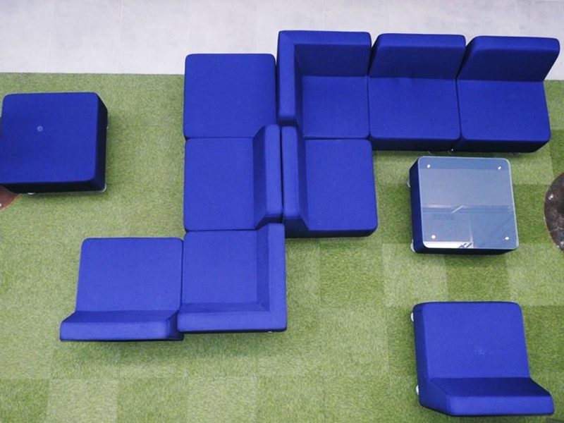 Niebieskie modułowe siedziska ustawione na zielonym dywanie z jednym przezroczystym siedziskiem.